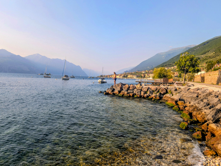 Abendstimmung am Gardasee — eine Person steht auf einem Felsvorsprung am Ufer, Segelboote liegen auf dem ruhigen Wasser, im Hintergrund grüne Berghänge im Dunst.