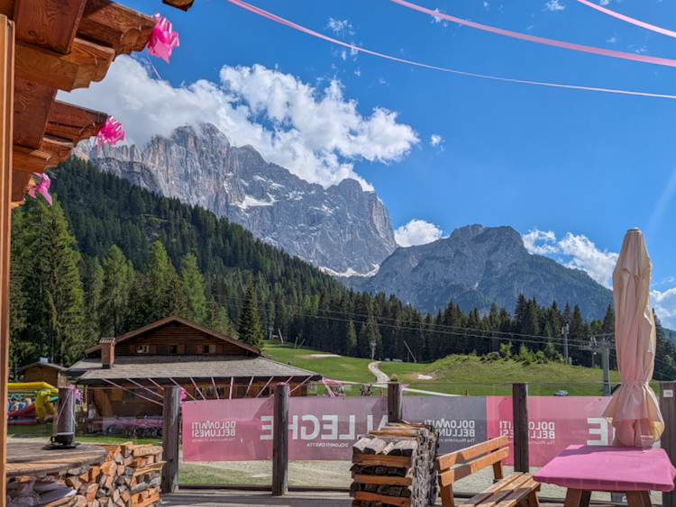 Blick von einer Holzhütte auf die Dolomiten in Alleghe — im Vordergrund rosa Giro-d'Italia-Dekoration und ein Banner mit der Aufschrift „Dolomiti Bellunesi", im Hintergrund mächtige Felsgipfel unter blauem Himmel.