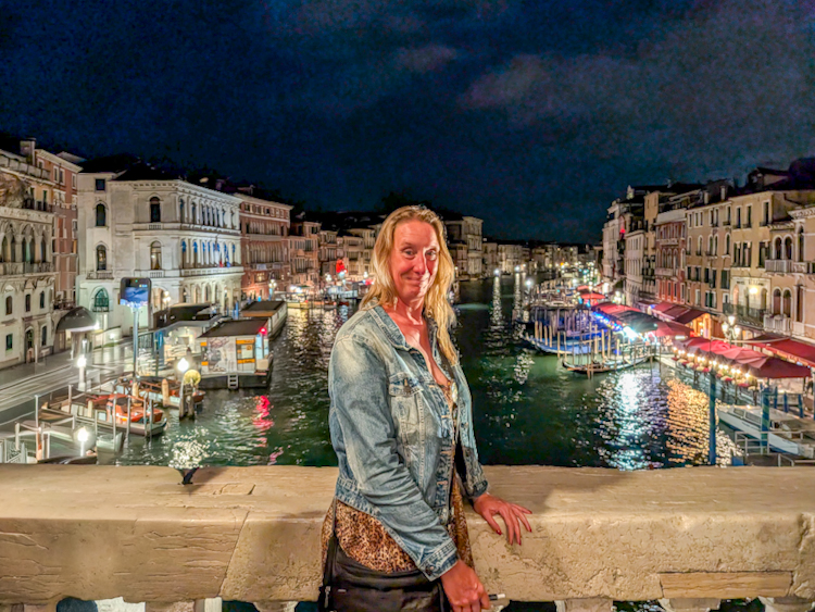 Sabine Schönberg steht abends auf der beleuchteten Rialtobrücke in Venedig, im Hintergrund der Canal Grande bei Nacht mit glitzernden Lichtern.