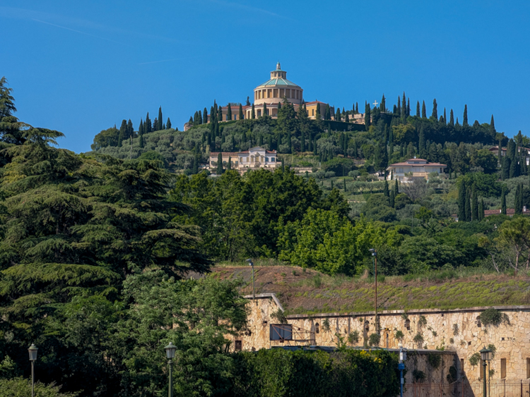 Blick auf einen bewaldeten Hügel in Verona mit der Basilika Madonna di Lourdes, umgeben von Zypressen und mediterraner Vegetation unter strahlend blauem Himmel.