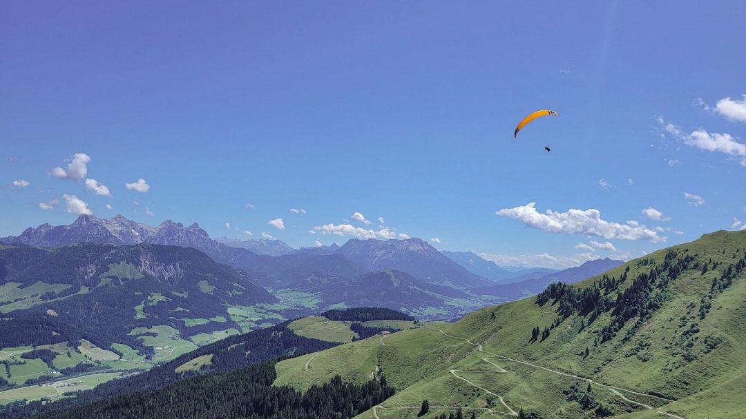 Panoramablick beim Wandern auf dem Koasa Trail: grüne Almhänge, weite Bergketten und ein Gleitschirm am blauen Himmel