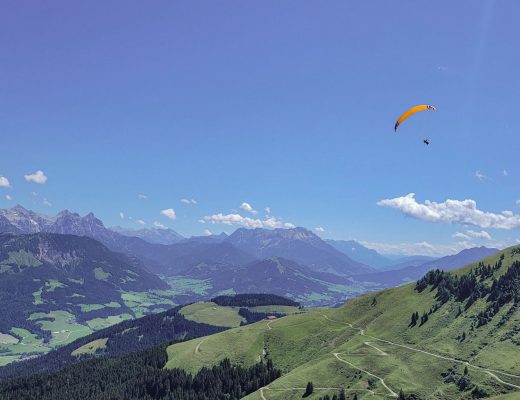 Panoramablick beim Wandern auf dem Koasa Trail: grüne Almhänge, weite Bergketten und ein Gleitschirm am blauen Himmel