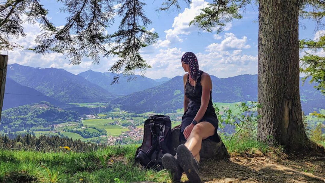Sabine Schönberg sitzt mit Wanderrucksack unter einem Baum und blickt über ein grünes Tal auf die Berge.